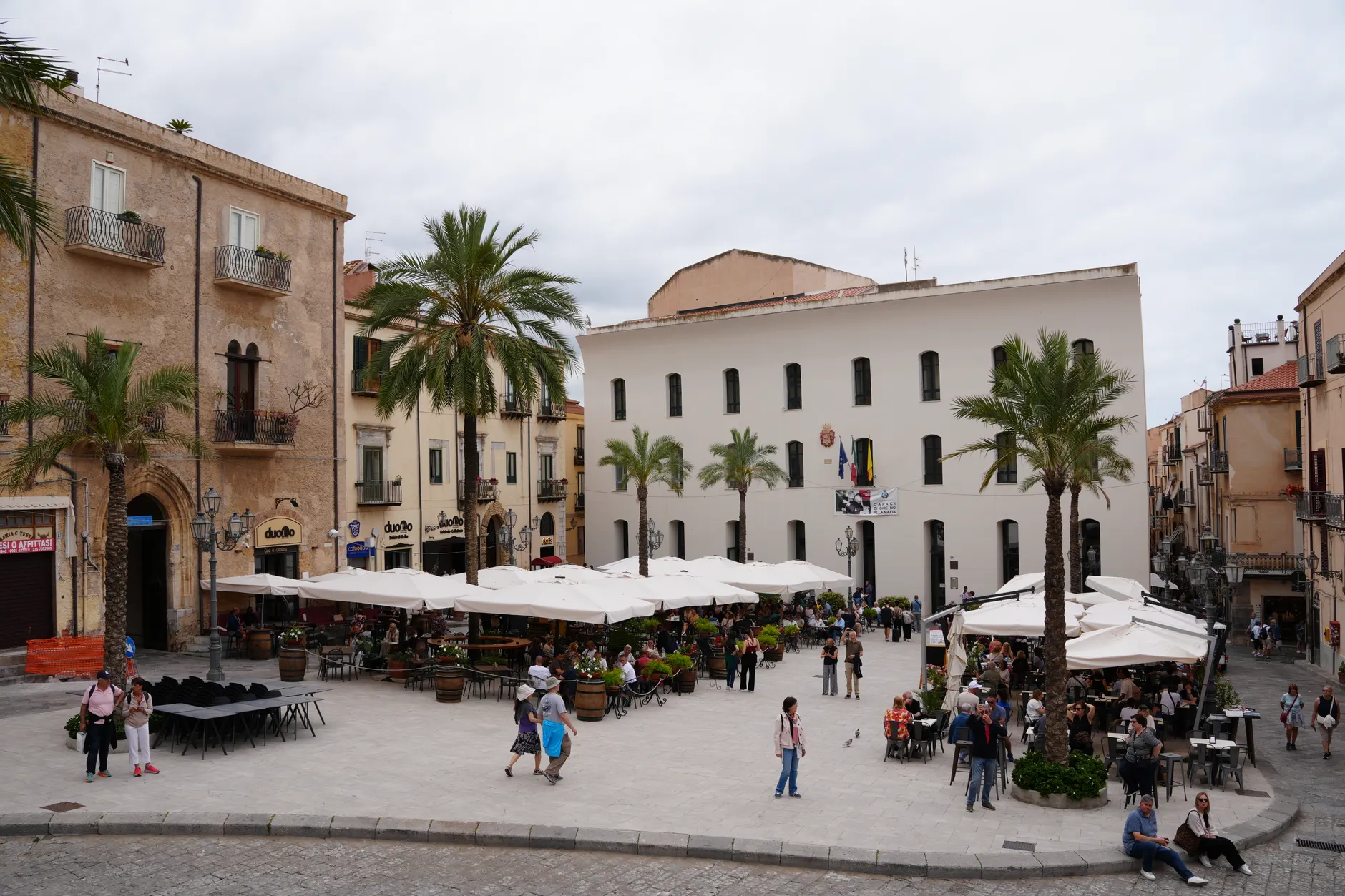 Busy Piazza Del Duomo in Cefalù, Italy, bustling with people and cafes under a cloudy sky.